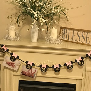 May include: A decorative banner with Minnie Mouse-themed cutouts, featuring numbers 1 through 6, and pink bows. The banner includes two photos of a baby. The banner is strung across a fireplace mantel, with a vase of flowers and candles.