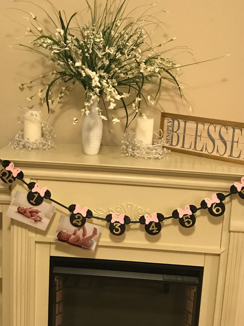 May include: A decorative banner with Minnie Mouse-themed cutouts, featuring numbers 1 through 6, and pink bows. The banner includes two photos of a baby. The banner is strung across a fireplace mantel, with a vase of flowers and candles.