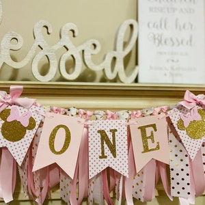 May include: A pink and gold "ONE" banner with Minnie Mouse silhouettes. The banner features pink pennants with gold glitter letters and polka dots, accented with pink ribbons and bows. A wooden "Blessed" sign is in the background.