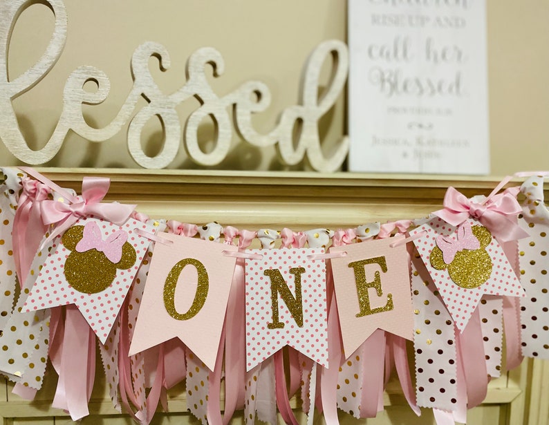 May include: A pink and gold "ONE" banner with Minnie Mouse silhouettes. The banner features pink pennants with gold glitter letters and polka dots, accented with pink ribbons and bows. A wooden "Blessed" sign is in the background.