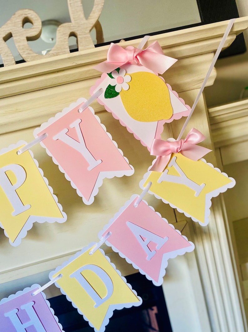 May include: A yellow and pink banner with the words "Happy Birthday" in white letters. The banner is decorated with pink bows and a lemon with a white flower.