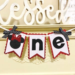 May include: A decorative banner spelling the word one with three white pennants featuring red glitter scalloped edges. The first pennant displays a black glitter mouse silhouette with a red bow, while the others show black letters n and e.