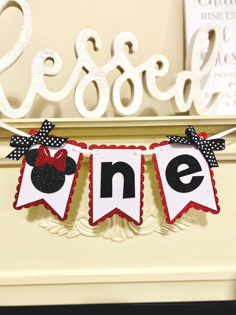 May include: A decorative banner spelling the word one with three white pennants featuring red glitter scalloped edges. The first pennant displays a black glitter mouse silhouette with a red bow, while the others show black letters n and e.