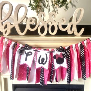 May include: A festive banner with pink, white, and black polka dot fabric strips. The banner features three white scalloped pennants with the letters "Two" and a Minnie Mouse silhouette. Wooden "Blessed" sign in the background.