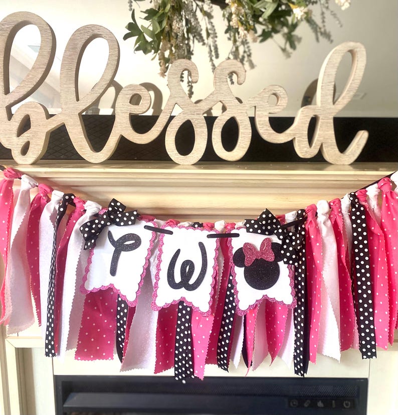 May include: A festive banner with pink, white, and black polka dot fabric strips. The banner features three white scalloped pennants with the letters "Two" and a Minnie Mouse silhouette. Wooden "Blessed" sign in the background.