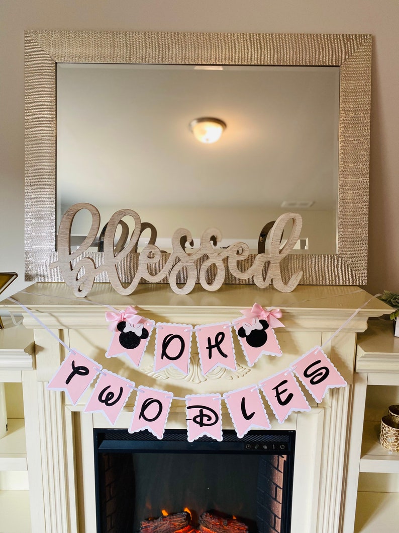 May include: Pink and white banner with the words "Oh Two D's" and black Mickey Mouse ears on each end. The banner is hanging above a fireplace.