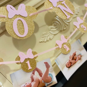 May include: A gold glitter Minnie Mouse themed banner with pink bows and numbers 0, 1, 2, and 3. The banner is strung with pink ribbon and has photos of a baby girl attached.