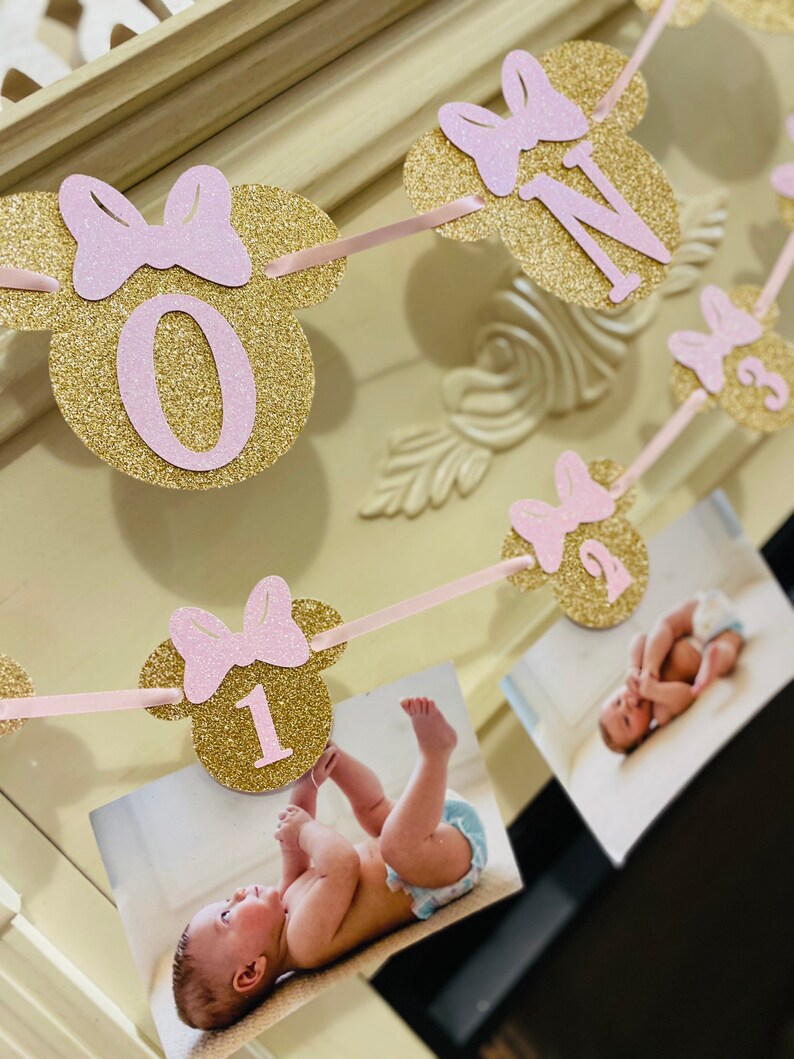 May include: A gold glitter Minnie Mouse themed banner with pink bows and numbers 0, 1, 2, and 3. The banner is strung with pink ribbon and has photos of a baby girl attached.
