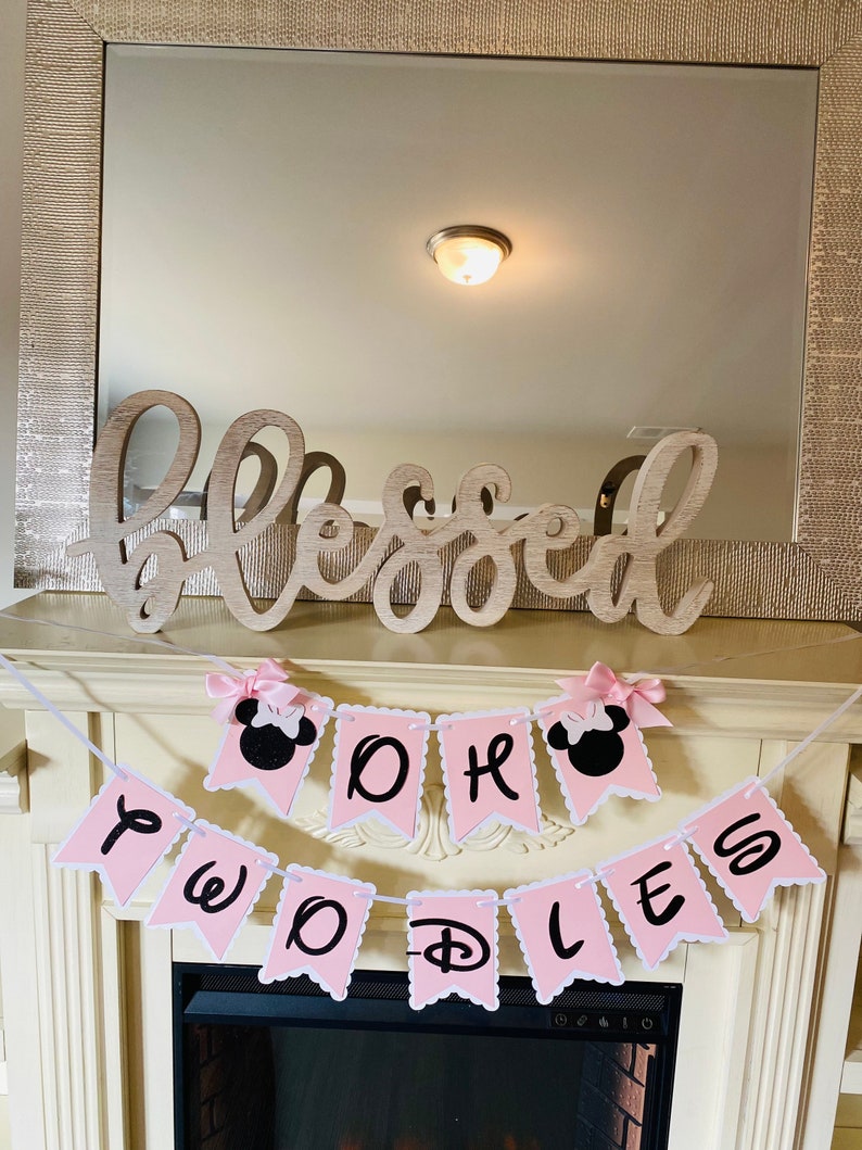 May include: Pink and black banner with the words "Twoodles Oh" and a Mickey Mouse head on each end. The banner is hanging from a fireplace mantel.