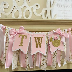 May include: A pink and gold banner with the word "TWO" in gold glitter letters. The banner has pink and white ribbons with gold polka dots and pink bows. A Minnie Mouse silhouette is on the right side.