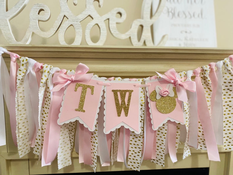 May include: A pink and gold banner with the word "TWO" in gold glitter letters. The banner has pink and white ribbons with gold polka dots and pink bows. A Minnie Mouse silhouette is on the right side.