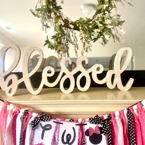 May include: A wooden "Blessed" sign sits above a banner with pink, white, and black polka dot fabric strips. The banner has three white pennants with the letters "P", "W", and a Minnie Mouse silhouette. A floral wreath hangs above.