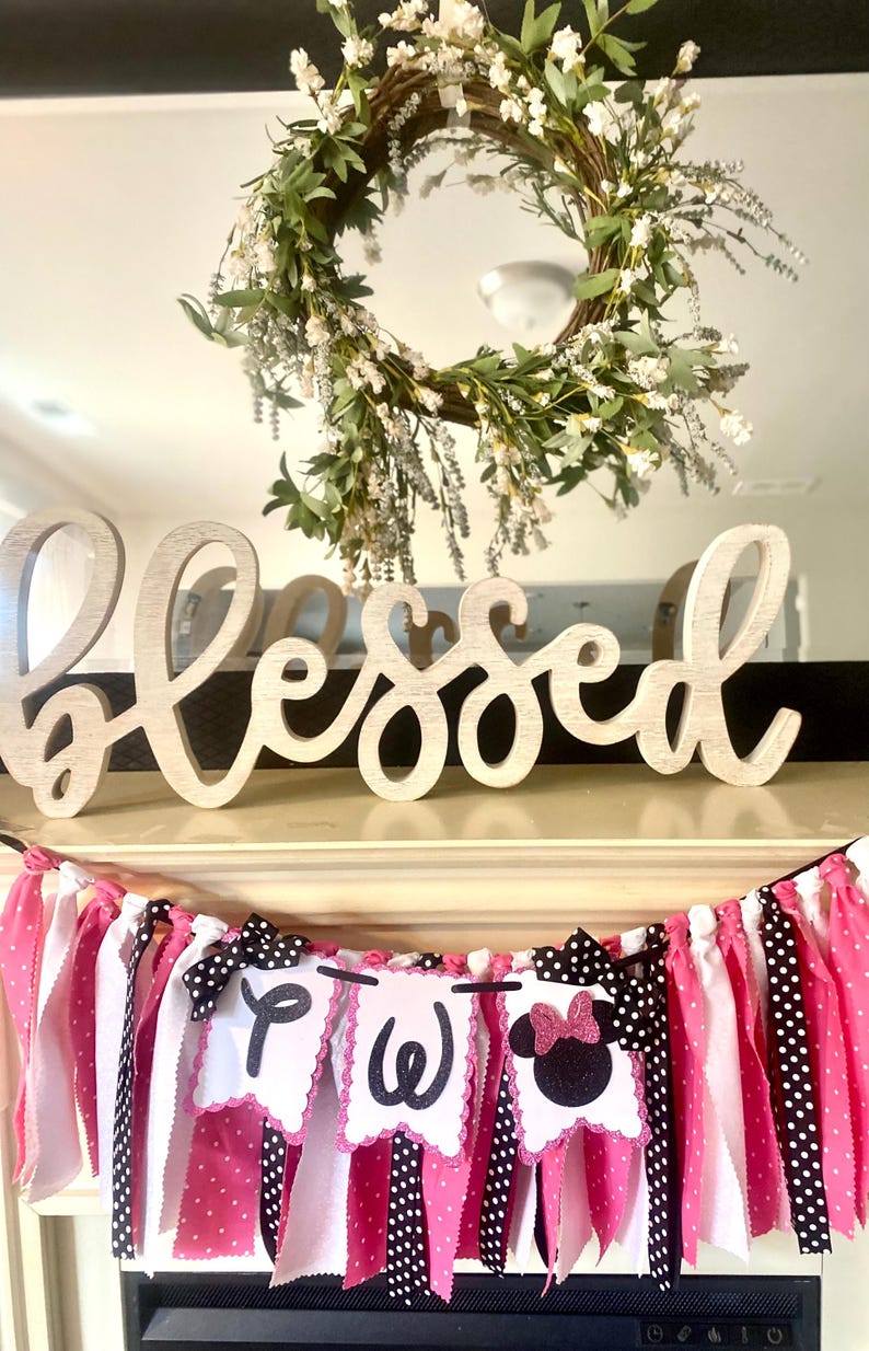 May include: A wooden "Blessed" sign sits above a banner with pink, white, and black polka dot fabric strips. The banner has three white pennants with the letters "P", "W", and a Minnie Mouse silhouette. A floral wreath hangs above.