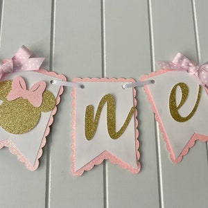 Puede incluir: Pancarta de "uno" con purpurina rosa y dorada con una cabeza de Minnie Mouse en el lado izquierdo. La pancarta est&aacute; decorada con lazos de lunares rosa y blanco.