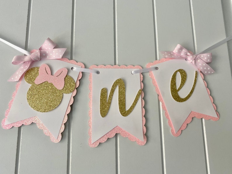 Puede incluir: Pancarta de "uno" con purpurina rosa y dorada con una cabeza de Minnie Mouse en el lado izquierdo. La pancarta est&aacute; decorada con lazos de lunares rosa y blanco.