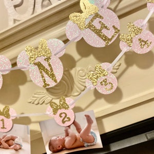 Op de afbeelding: Roze en gouden glitter Minnie Mouse-banner met het woord "ONE" en de nummers 1, 2, 3 en 4. De banner is aan een wit lint geregen en heeft een fotoplek voor elk nummer.
