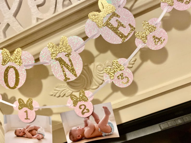 Op de afbeelding: Roze en gouden glitter Minnie Mouse-banner met het woord "ONE" en de nummers 1, 2, 3 en 4. De banner is aan een wit lint geregen en heeft een fotoplek voor elk nummer.