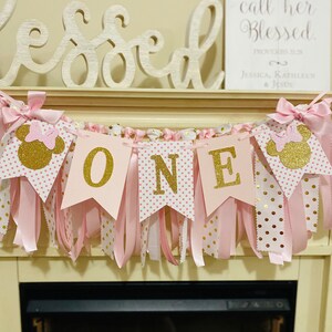 May include: A pink and gold "ONE" banner with Minnie Mouse silhouettes and bows. The banner has pink ribbons and polka dot flags. The word "Blessed" is above the banner.