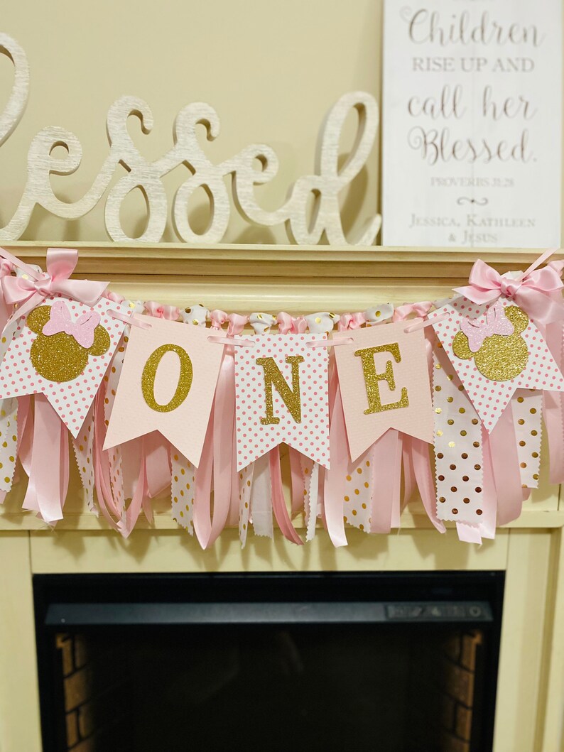 May include: A pink and gold "ONE" banner with Minnie Mouse silhouettes and bows. The banner has pink ribbons and polka dot flags. The word "Blessed" is above the banner.