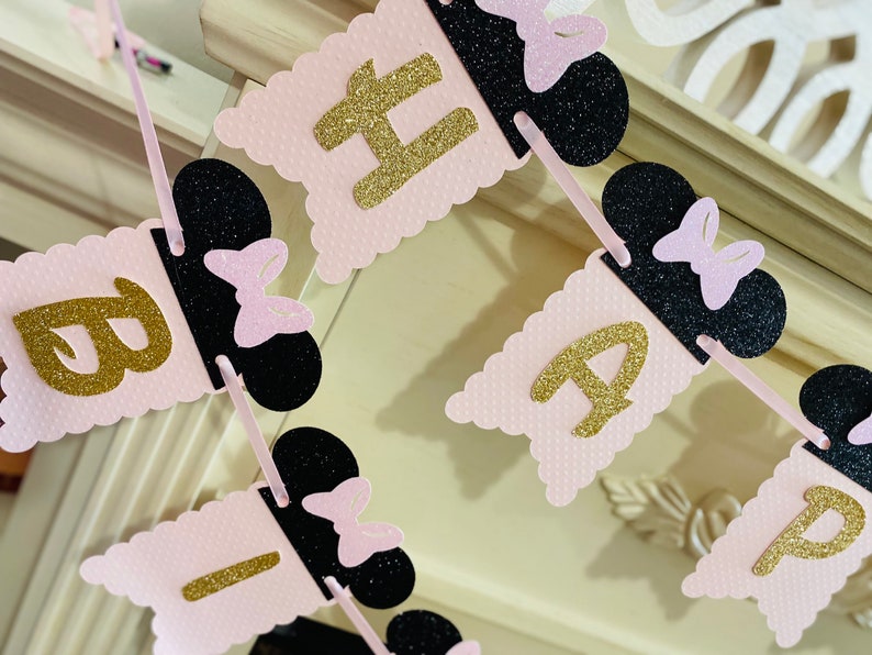 May include: Pink and black banner with gold glitter letters spelling "Happy Birthday". Each letter is on a separate flag with a black Mickey Mouse ear and pink bow.