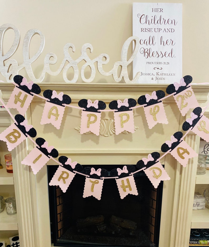 May include: Pink and black Happy 2nd Birthday banner with Minnie Mouse ears and bows. The banner is strung up over a fireplace.