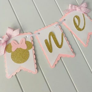 Puede incluir: Bandera de brillo rosa y dorado con la palabra "one" y una cabeza de Minnie Mouse con un lazo rosa.