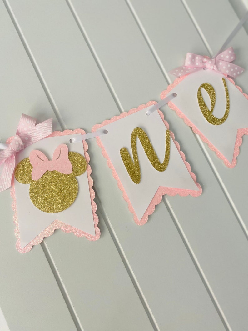 Puede incluir: Bandera de brillo rosa y dorado con la palabra "one" y una cabeza de Minnie Mouse con un lazo rosa.