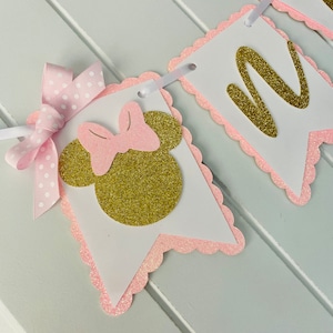 Puede incluir: Bandera de Minnie Mouse en rosa y dorado con la letra "M" en purpurina dorada. La bandera est&aacute; hecha de cartulina y tiene un borde festoneado.