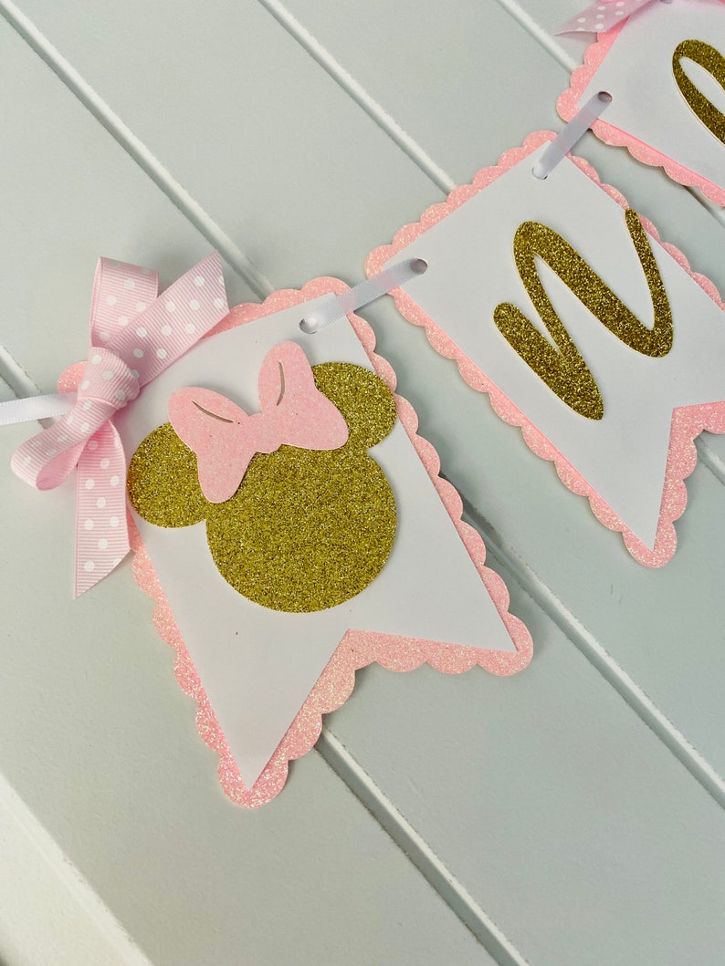 Puede incluir: Bandera de Minnie Mouse en rosa y dorado con la letra "M" en purpurina dorada. La bandera est&aacute; hecha de cartulina y tiene un borde festoneado.