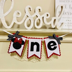 May include: A decorative banner featuring three white pennants with scalloped red glitter edges. The first pennant displays a black glitter Minnie Mouse silhouette with a red bow. The remaining two pennants feature the black letters n and e.