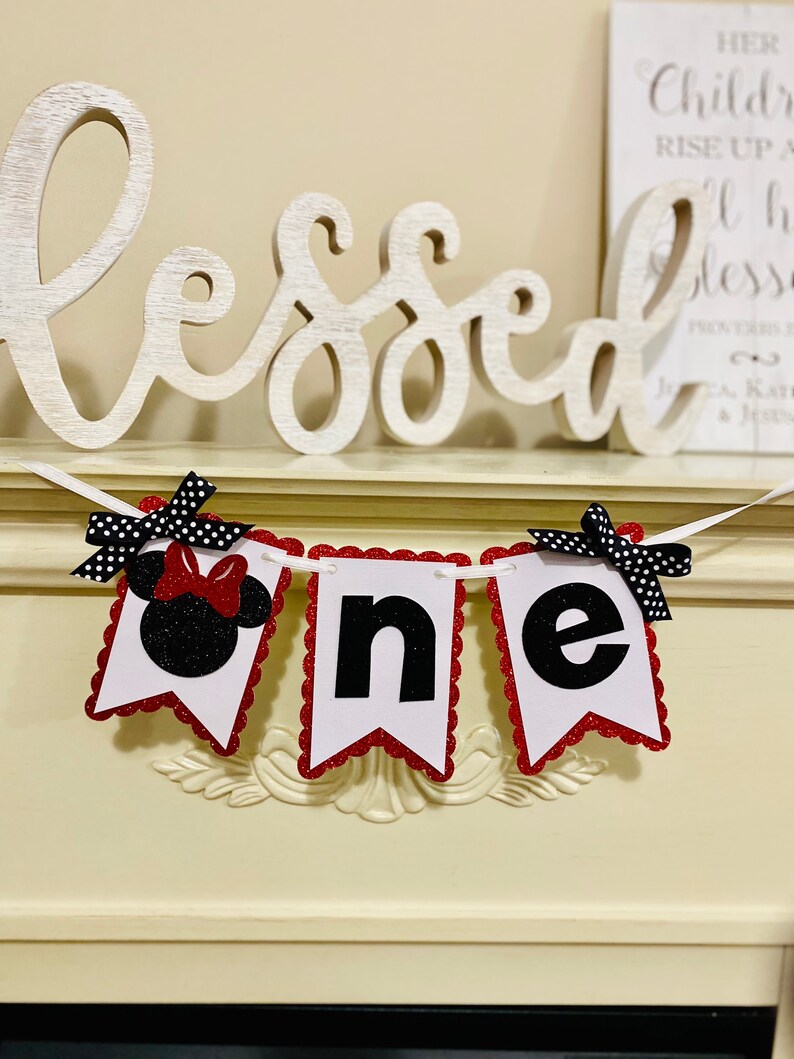 May include: A decorative banner featuring three white pennants with scalloped red glitter edges. The first pennant displays a black glitter Minnie Mouse silhouette with a red bow. The remaining two pennants feature the black letters n and e.