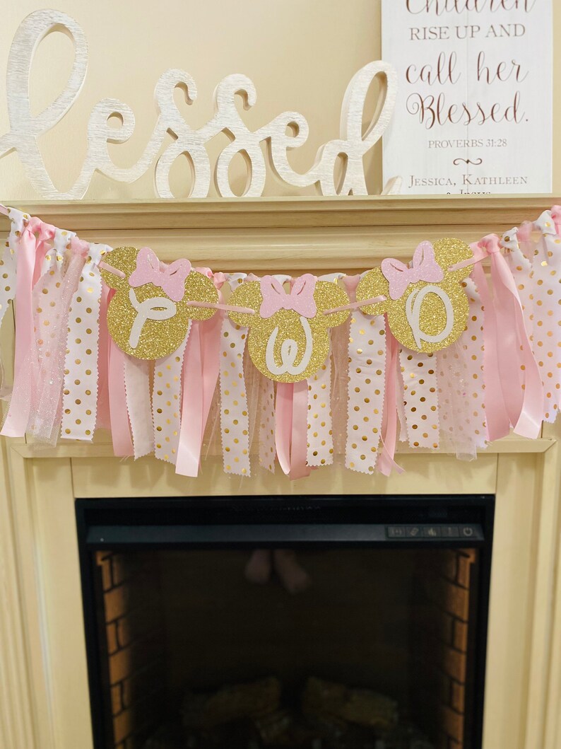 May include: A decorative banner with gold glitter Minnie Mouse heads spelling out a name, with pink bows and ribbons. The banner is strung with pink and gold polka dot fabric strips. Above the banner is a wooden sign that says "Blessed".