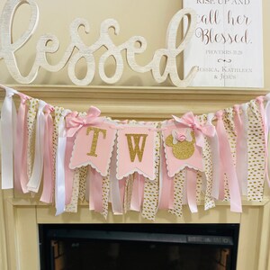 May include: A pink and gold banner with the letters "TW" in gold glitter, accented with pink bows and ribbons. The banner is decorated with white and gold polka dot ribbons. A wooden sign above reads "Her Children Rise Up and call her Blessed."