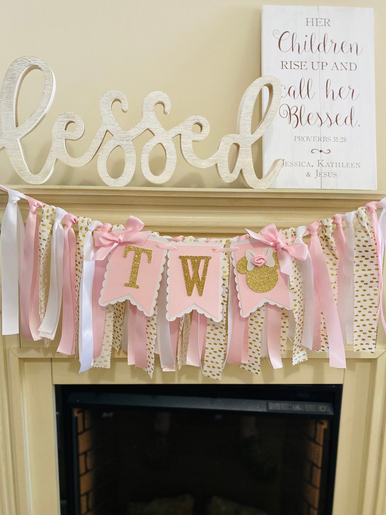 May include: A pink and gold banner with the letters "TW" in gold glitter, accented with pink bows and ribbons. The banner is decorated with white and gold polka dot ribbons. A wooden sign above reads "Her Children Rise Up and call her Blessed."