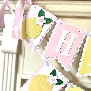 May include: A decorative banner with pink, white, and yellow scalloped edges. The banner features lemon and flower cutouts, with the letter "H" in white. A pink ribbon is tied at the top.