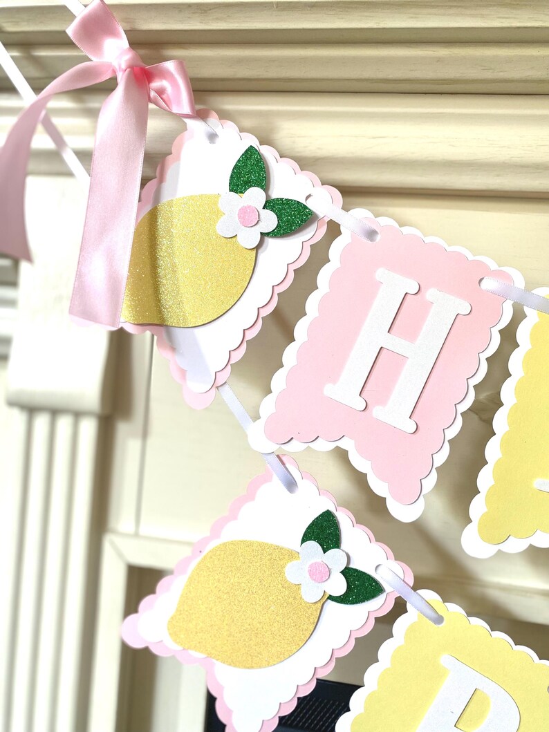 May include: A decorative banner with pink, white, and yellow scalloped edges. The banner features lemon and flower cutouts, with the letter "H" in white. A pink ribbon is tied at the top.