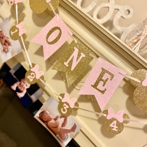 Puede incluir: Bandera de brillo rosa y dorado con la palabra "ONE" y orejas de Mickey Mouse con lazos rosas. La bandera tiene fotos de una ni&ntilde;a en cada secci&oacute;n.