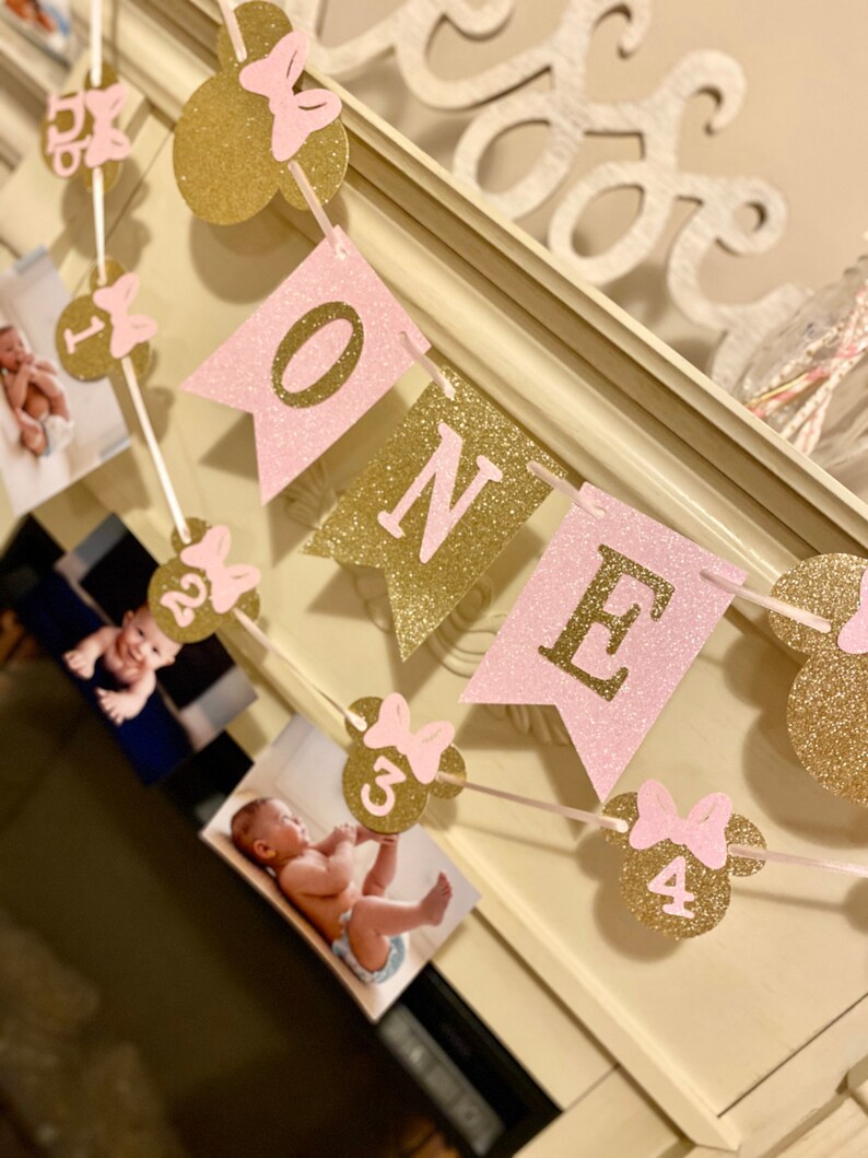 Puede incluir: Bandera de brillo rosa y dorado con la palabra "ONE" y orejas de Mickey Mouse con lazos rosas. La bandera tiene fotos de una ni&ntilde;a en cada secci&oacute;n.
