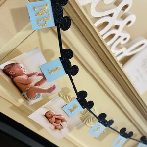 Pode incluir: Uma faixa azul e preta com orelhas do Mickey Mouse e n&uacute;meros de glitter dourado. A faixa &eacute; decorada com fotos de um beb&ecirc;. A faixa diz "Um".