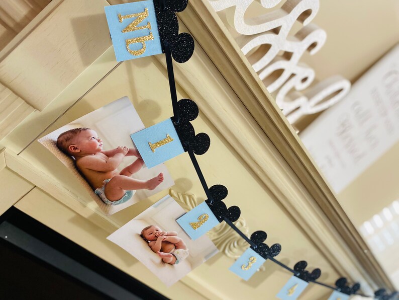 Pode incluir: Uma faixa azul e preta com orelhas do Mickey Mouse e n&uacute;meros de glitter dourado. A faixa &eacute; decorada com fotos de um beb&ecirc;. A faixa diz "Um".