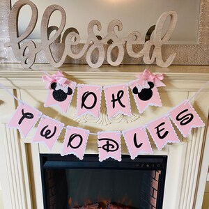 May include: Pink and white banner with black glitter letters that reads "One Two D-O-L-L-E-S". The banner is decorated with pink bows and black Mickey Mouse ears. The banner is hanging above a fireplace with a wooden sign that reads "Blessed".