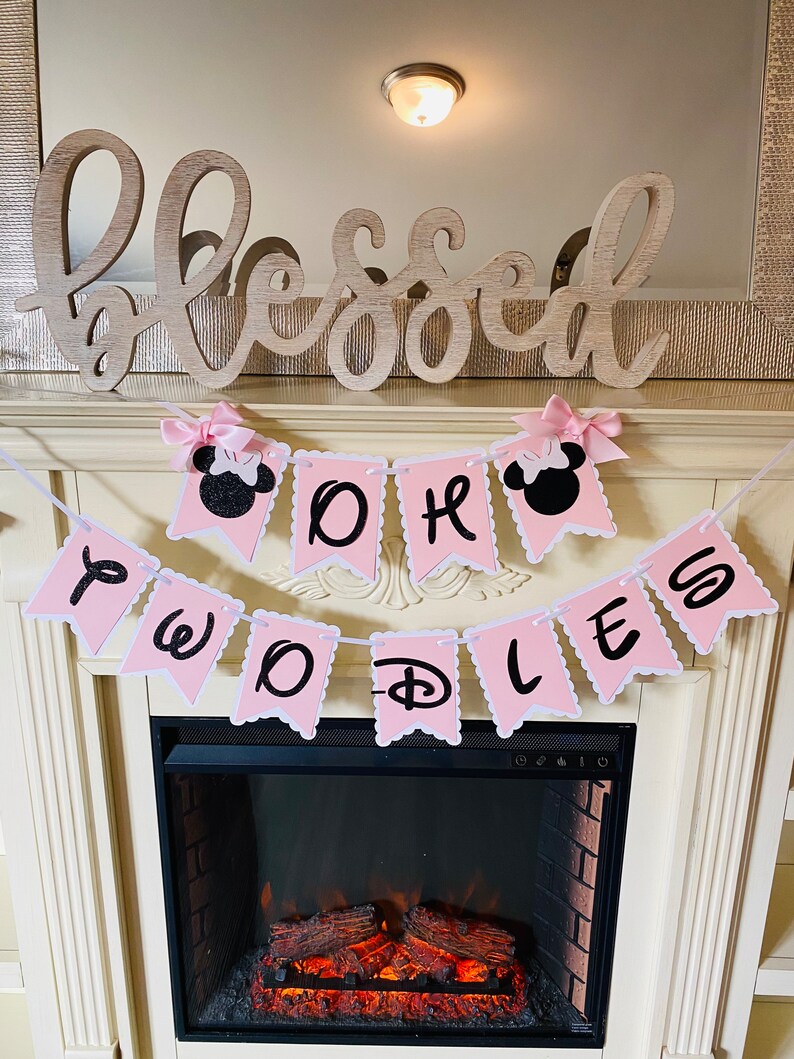 May include: Pink and white banner with black glitter letters that reads "One Two D-O-L-L-E-S". The banner is decorated with pink bows and black Mickey Mouse ears. The banner is hanging above a fireplace with a wooden sign that reads "Blessed".