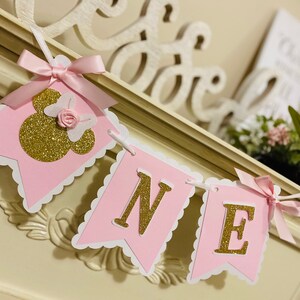 Puede incluir: Bandera rosa y dorada con la palabra "ONE" y una cabeza de Minnie Mouse de purpurina dorada con un lazo rosa.