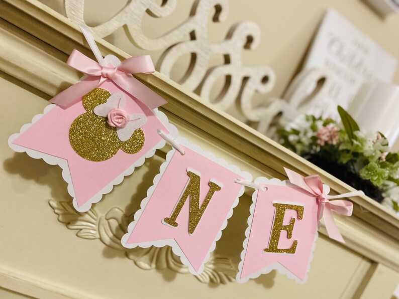 Puede incluir: Bandera rosa y dorada con la palabra "ONE" y una cabeza de Minnie Mouse de purpurina dorada con un lazo rosa.