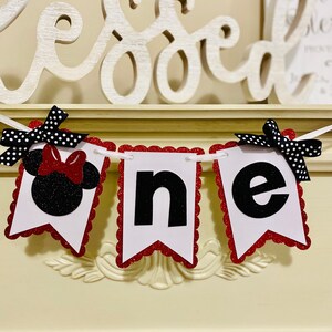 May include: A decorative banner spelling the word one, featuring three white scalloped-edge pennants with red glitter borders. The first pennant displays a black glitter mouse silhouette with a red bow, while the others show black letters.