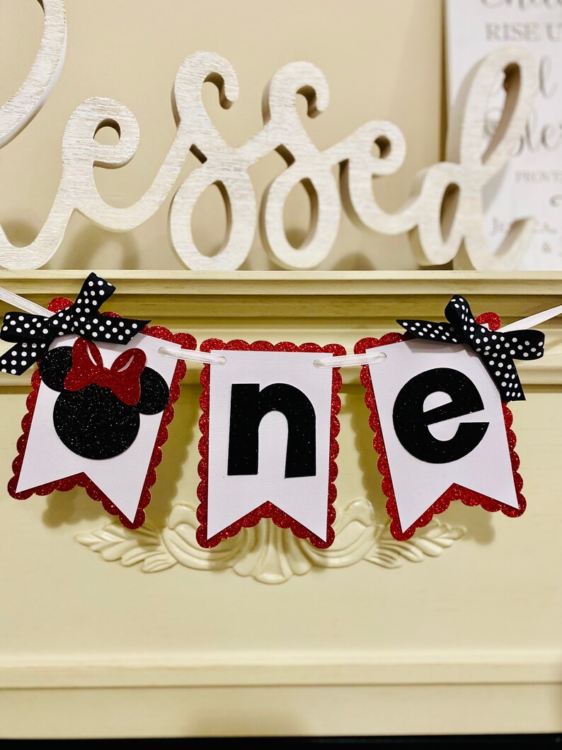 May include: A decorative banner spelling the word one, featuring three white scalloped-edge pennants with red glitter borders. The first pennant displays a black glitter mouse silhouette with a red bow, while the others show black letters.