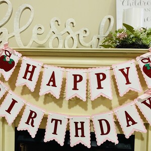 May include: A pink and white banner with red letters that reads "Happy Birthday". The banner has a scalloped edge and is decorated with red apples with green leaves and glitter.