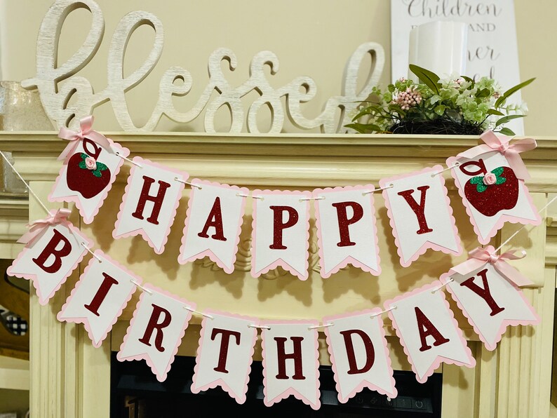 May include: A pink and white banner with red letters that reads "Happy Birthday". The banner has a scalloped edge and is decorated with red apples with green leaves and glitter.