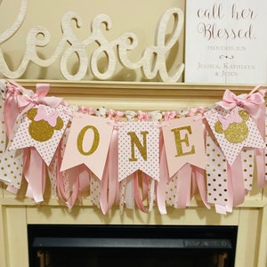 May include: A pink and gold "ONE" banner with Minnie Mouse silhouettes, ribbons, and polka dots. The banner is part of a first birthday party theme. The word "Blessed" is displayed above the banner.
