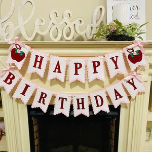 May include: Pink and white banner with red letters that reads "Happy Birthday". The banner has red apple cutouts with green leaves and glitter. The banner is hanging above a white fireplace.